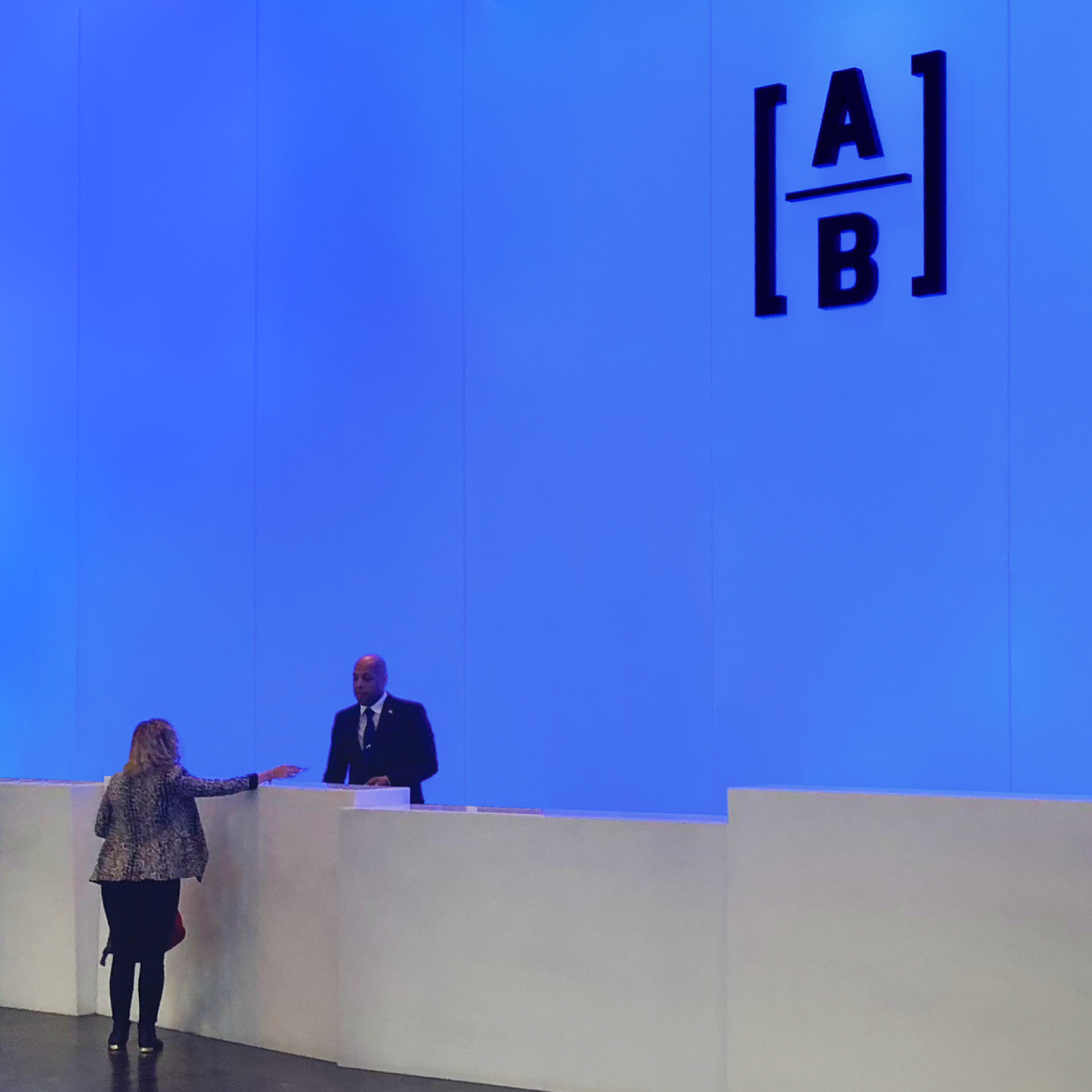 a woman hands her ID to a man behind a counter with a blue wall with the AB logo.