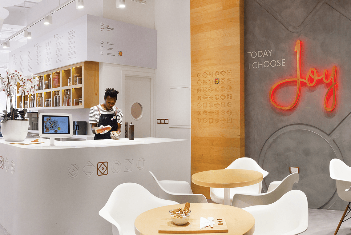 a person standing behind the counter serving yogurt in a room with a table and chairs.