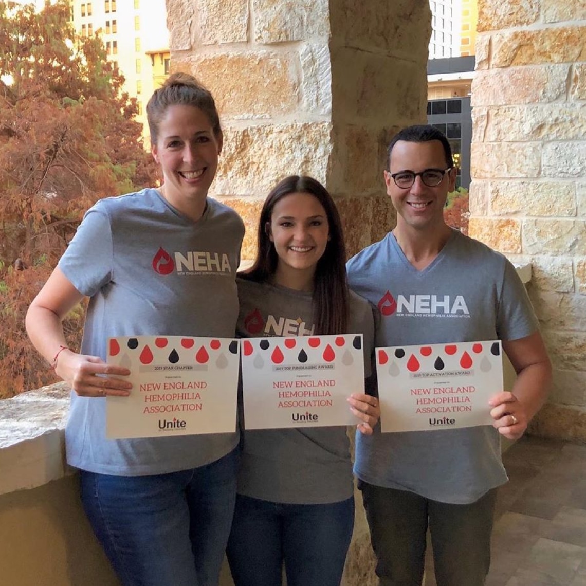 a group of three people, standing next to each other, displaying certificates and wearing T-shirts with the NEHA logo