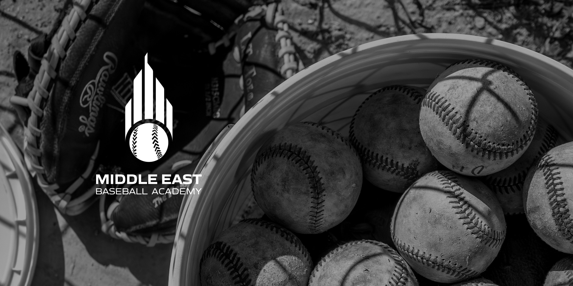 A black and white image of a basket of baseballs and gloves which showcases the black and white version of the Middle East Baseball Academy logo.