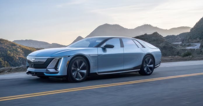 A sleek, silver electric car drives on a mountain road with hills and a clear sky in the background.