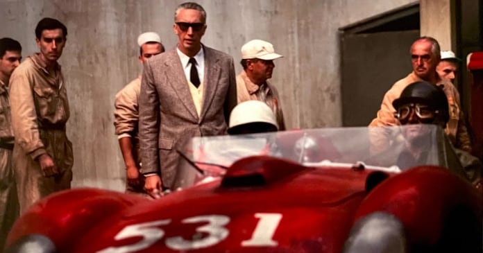 A group of men, some in work uniforms, stand around a red race car with the number 531; a man in a suit and sunglasses stands at the center.