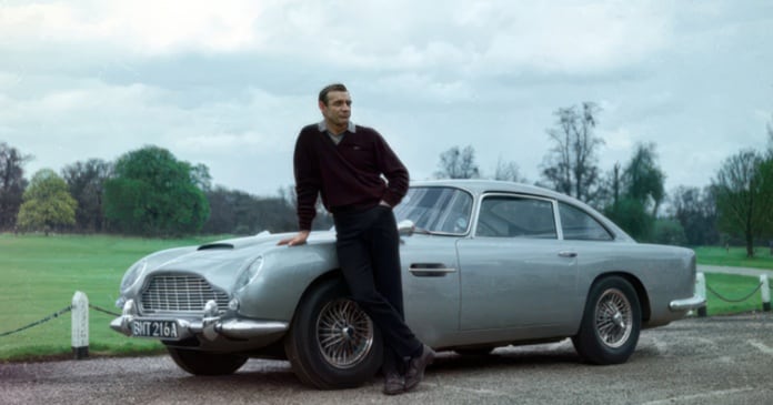 A man in dark clothing leans against a silver classic car parked on a road, with a grassy field and trees in the background.